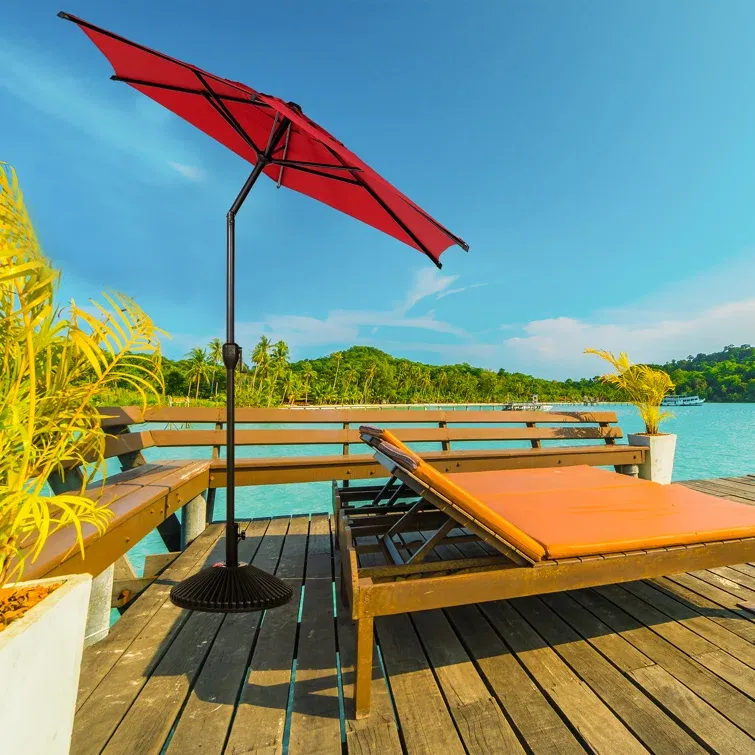 Two wooden sun loungers with orange cushions and a red umbrella on a wooden deck, overlooking a beachfront with clear skies.