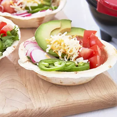A waffle bowl filled with avocado, cheese, tomato, onion, and jalapeño slices sits on a wooden surface.