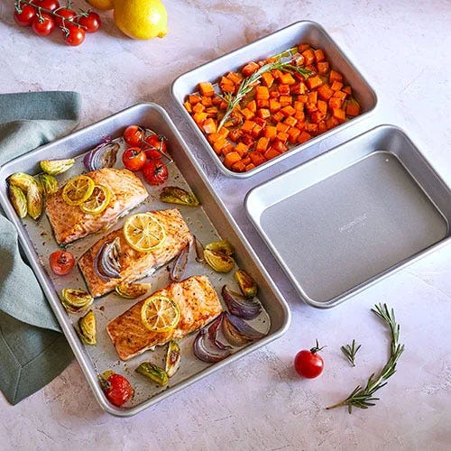 Stainless steel baking sheets with prepared salmon and vegetables, and a small container with diced sweet potatoes.
