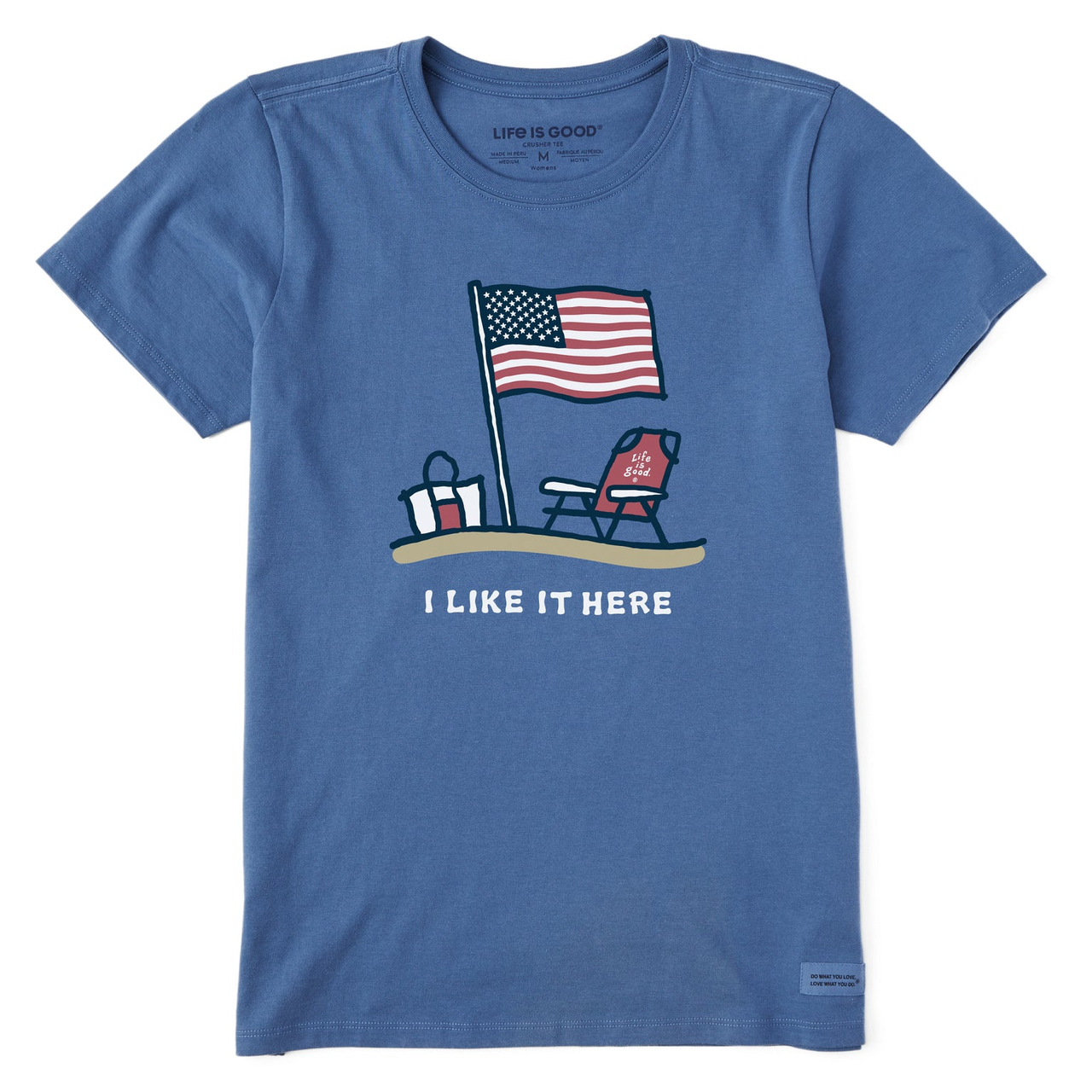 Blue t-shirt featuring an American flag, beach chair, beach bag, and the text \“I LIKE IT HERE.\“ The chair has \“Life is Good\“ written on it.
