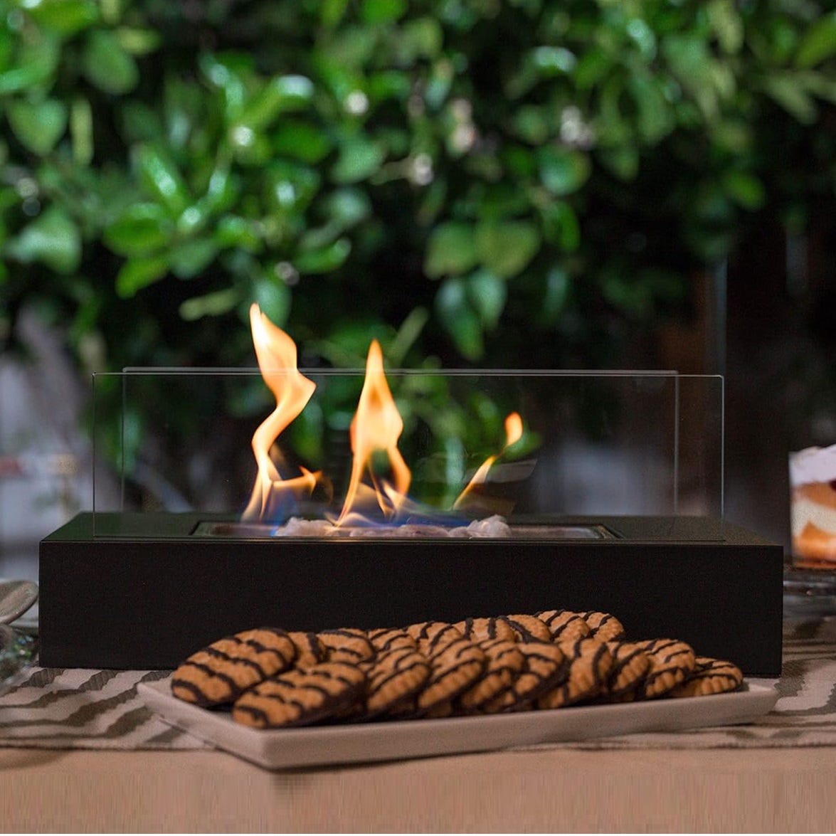 A tabletop bioethanol fireplace with visible flames and a plate of striped cookies in the foreground.