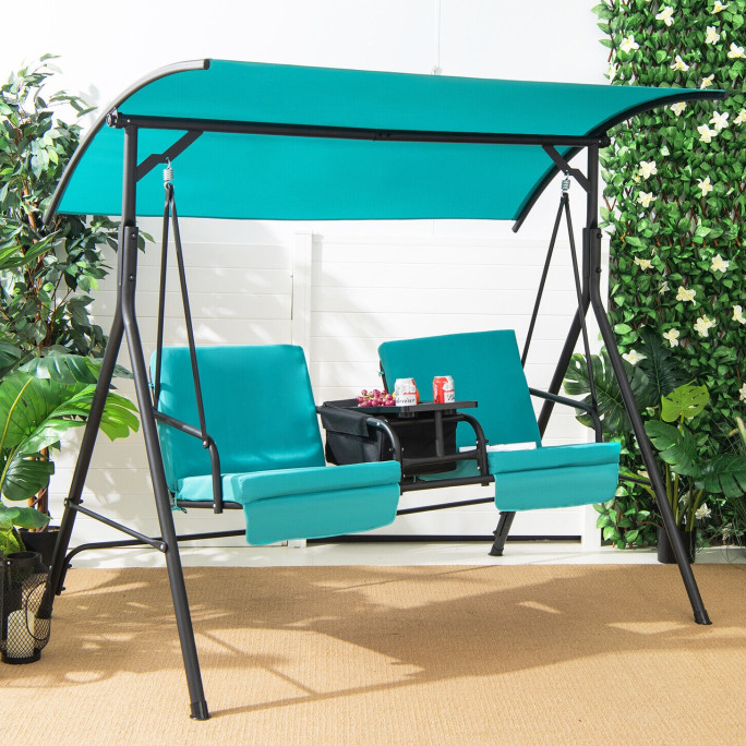A turquoise two-seater canopy swing with cushioned seats features a central black cooler table, housing two drinks and snacks, set on a light brown mat surrounded by greenery.
