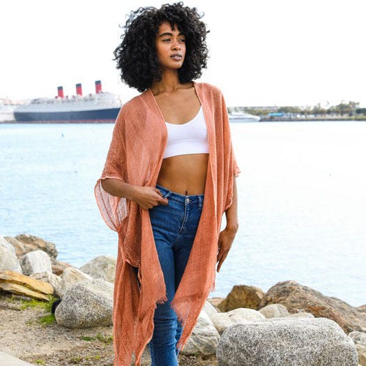 A woman wearing a white crop top, blue jeans, and a draped peach shawl by a waterfront.