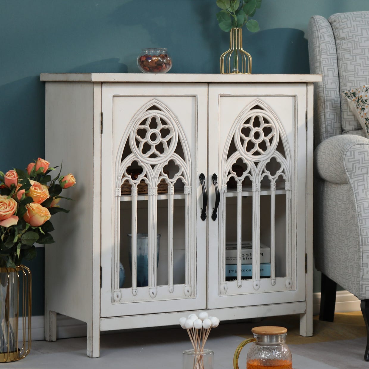 Vintage-style white wooden cabinet with gothic arch glass doors, placed in a room alongside decorative items.