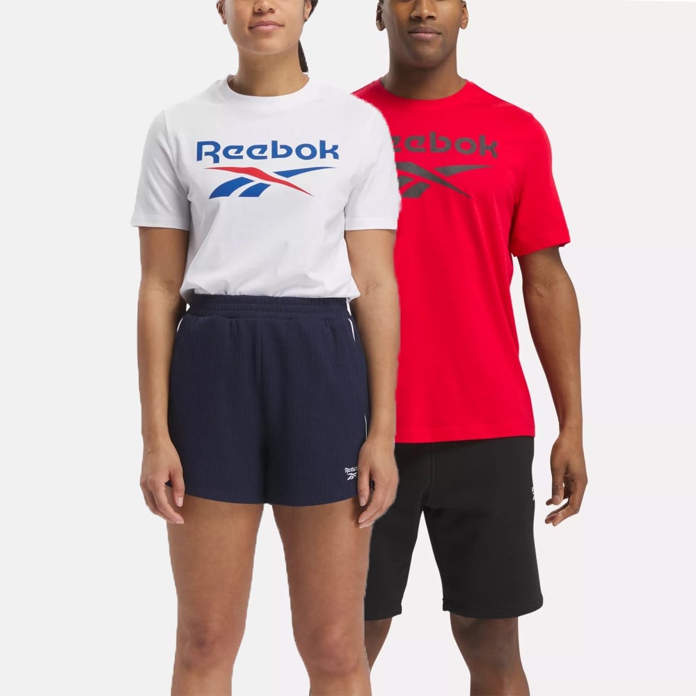 Two people wearing Reebok branded clothing; the person on the left in a white t-shirt with a blue logo and navy shorts, and the person on the right in a red t-shirt with a red logo and black shorts.