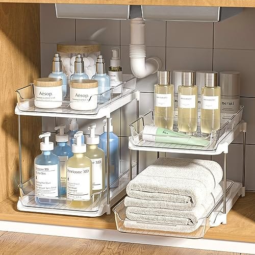Two tiered, sliding clear plastic cabinet organizers are shown storing various bottles and towels under a sink.