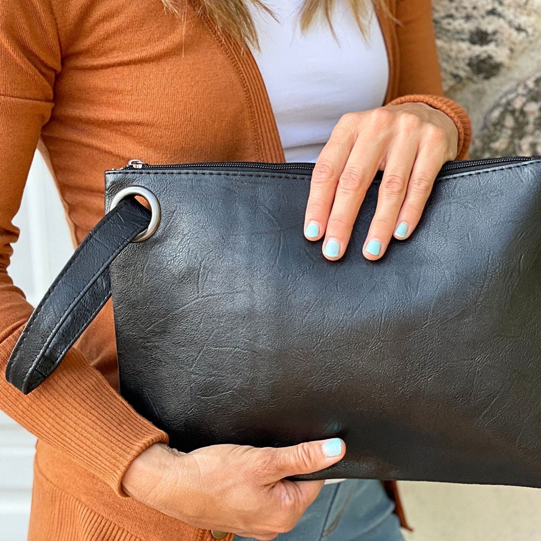 A person is holding a black leather clutch with a wrist strap and a zipper closure; the person has blue nail polish.