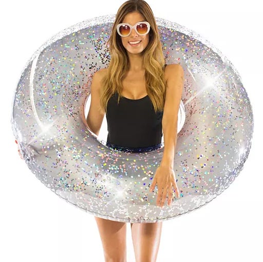 A person is holding a large, transparent, glitter-filled inflatable pool float. They are wearing white-rimmed sunglasses and a black swimsuit.