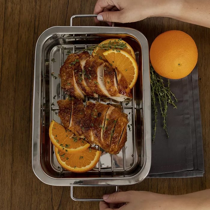 A sliced roasted chicken with orange slices on a metal serving tray, with fresh thyme, an orange, and a cloth napkin beside it.