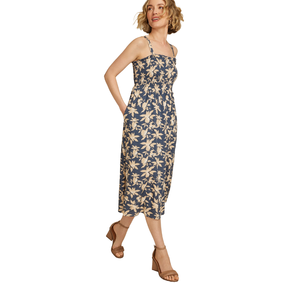 A woman is wearing a sleeveless knee-length dress with thin straps and a smocked bodice, featuring a blue background and a light floral print. She pairs it with tan wedge sandals.