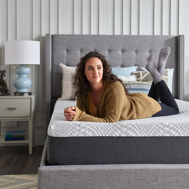 A woman lounging on a gray upholstered bed with a blue lamp and white nightstand in the background.