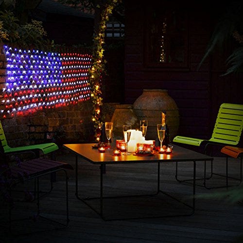 A lit LED American flag hangs in a dark setting, bright in red, white, and blue, next to some outdoor furniture and candles on a table.