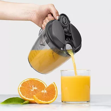 A person is pouring orange juice from a clear, gray lidded NutriBullet juicer pitcher into a glass, with halved oranges beside it.