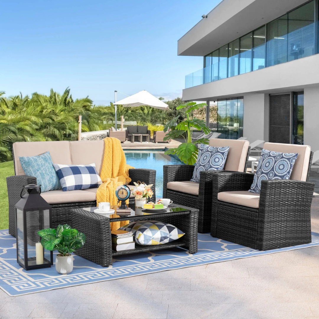 4-piece patio set includes a cushioned loveseat, two cushioned armchairs, and a glass-top coffee table. The furniture features dark wicker with beige cushions and is accessorized with decorative pillows, a throw blanket, and outdoor accents.