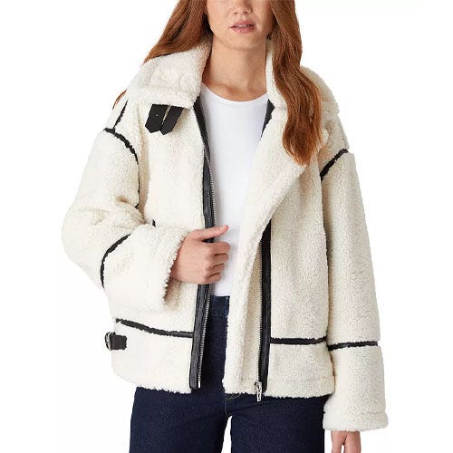 A woman is wearing a white sherpa jacket with black accents, featuring a front zipper and adjustable side buckles.