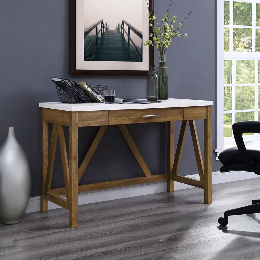 The Walker Edison Rustic Console Table features a modern design with a white tabletop, natural wood frame, and a single drawer. It includes a decorative vase, books, and a picture frame on top, set against a room with a gray wall and large window.