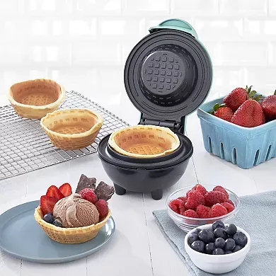 A Dash Mini Waffle Bowl Maker is open, displaying a freshly made waffle bowl inside, with finished bowls and berries on the countertop.