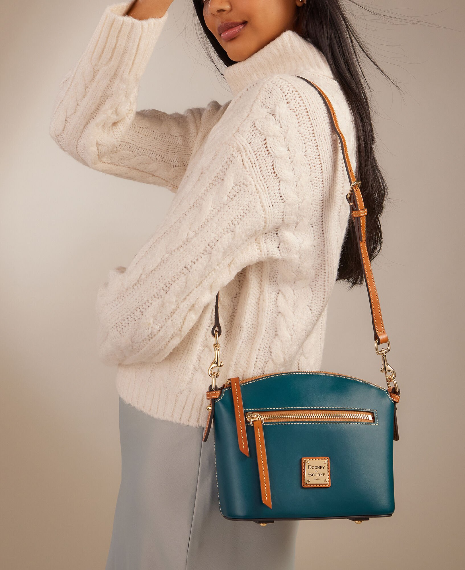 A woman is wearing a white cable knit sweater and carrying a teal crossbody bag with tan accents.