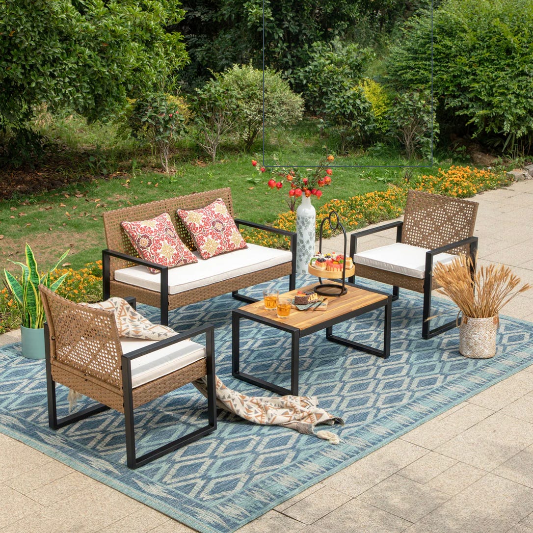 A patio furniture set with a two-seater sofa, two armchairs, and a coffee table, placed on an outdoor rug amidst a garden setting.