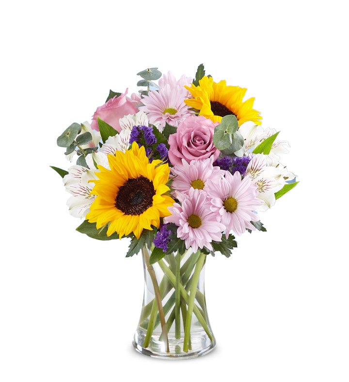 A vibrant bouquet featuring sunflowers, pink roses, pale pink daisies, white alstroemerias, and purple accent flowers, arranged with green foliage in a clear glass vase.