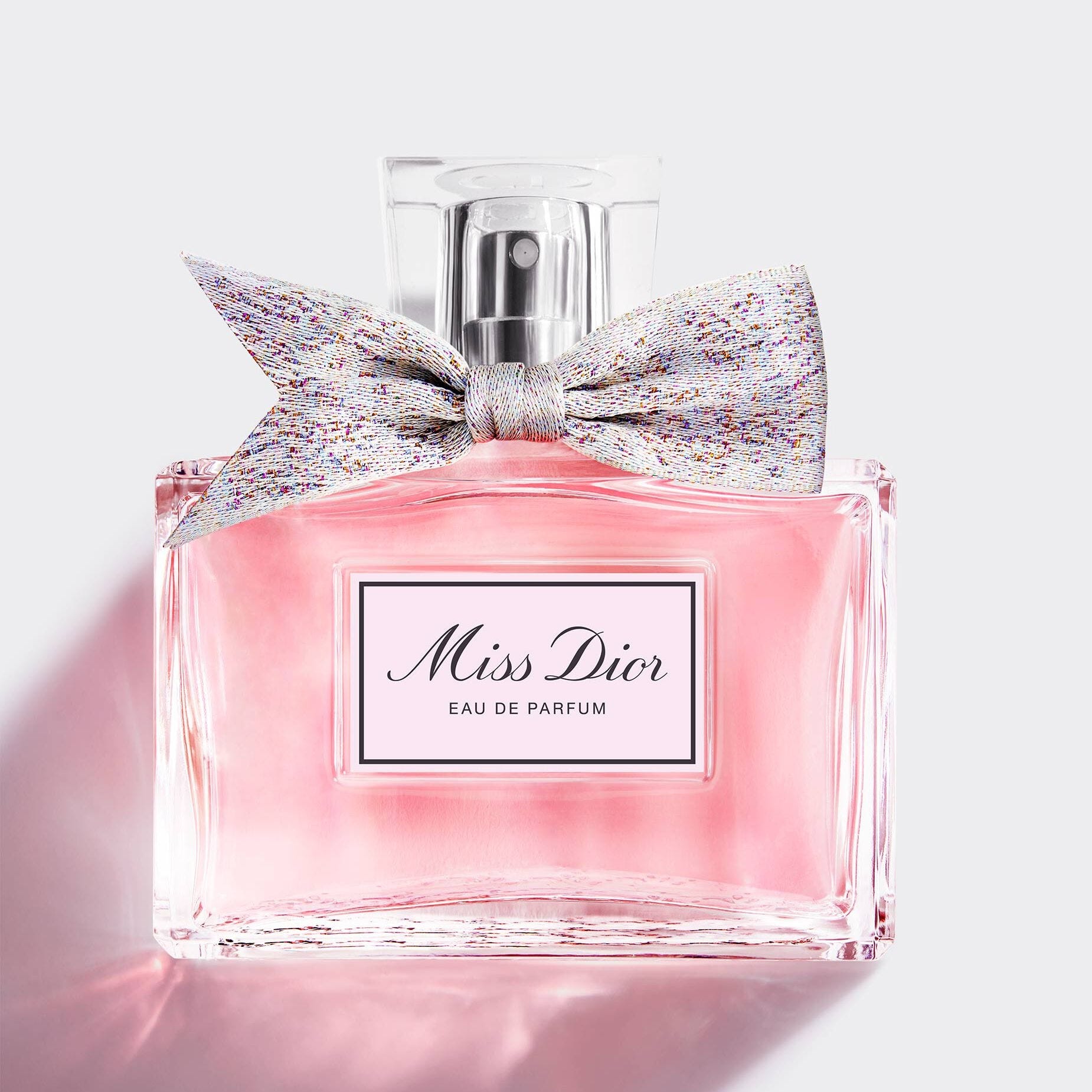 A square-shaped bottle of Miss Dior Eau de Parfum with pink liquid and a multi-colored bow.