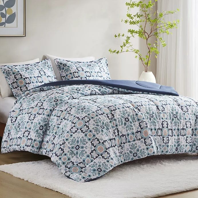 A bed is shown with a patterned comforter and two matching pillows, featuring a floral and geometric design in blue, grey, and white. A potted plant and framed artwork are in the background.