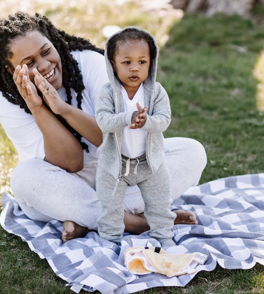 A woman and a toddler on a checkered blanket; the child is wearing a grey hoodie and pants set.