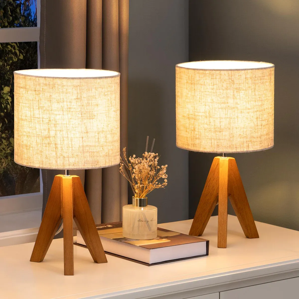 Two wooden tripod table lamps with cylindrical fabric shades are on a white surface, accompanied by a decorative bottle and a book.