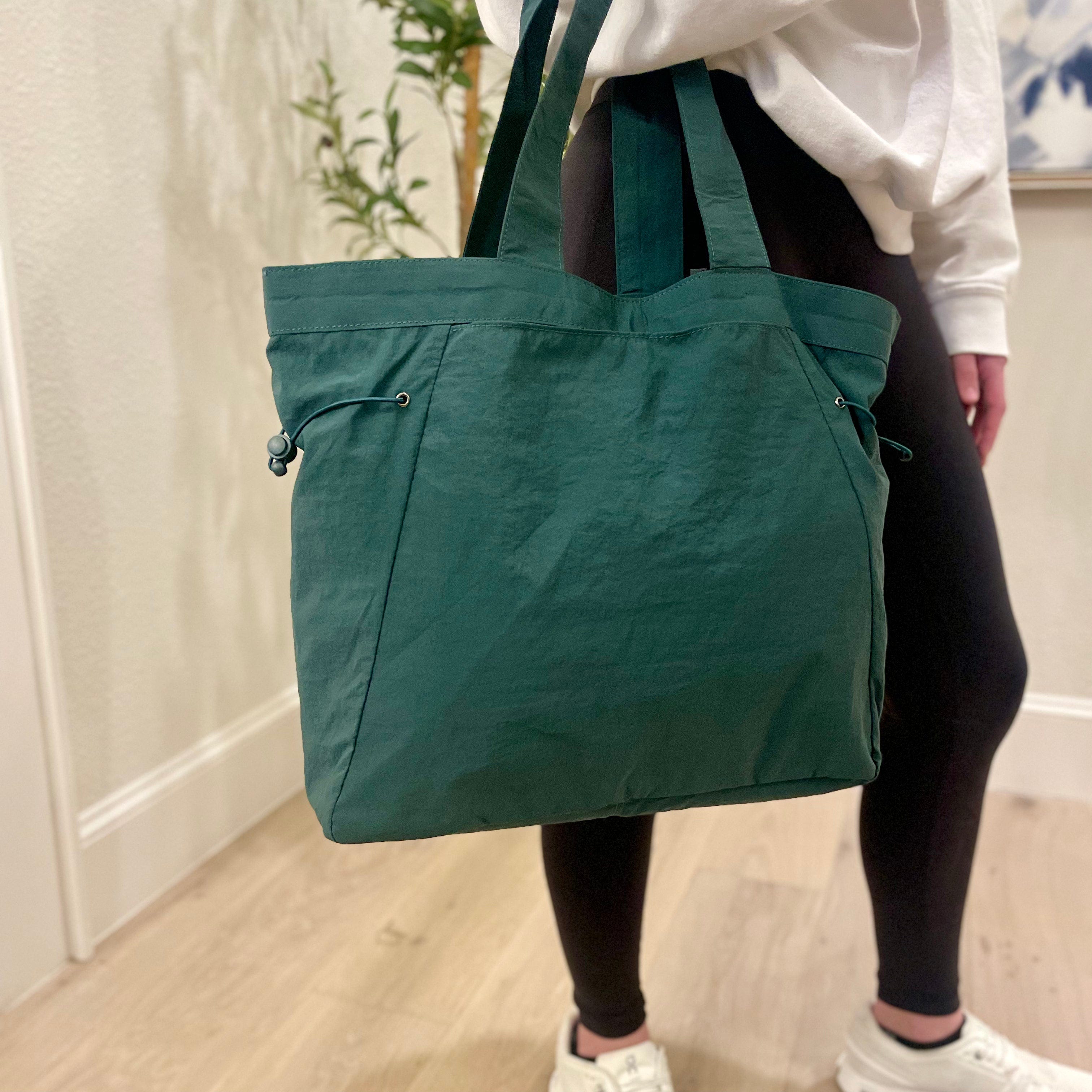 A person is holding a green cinch tote bag with long straps; the bag features an outer zip pocket and a drawstring on one side.