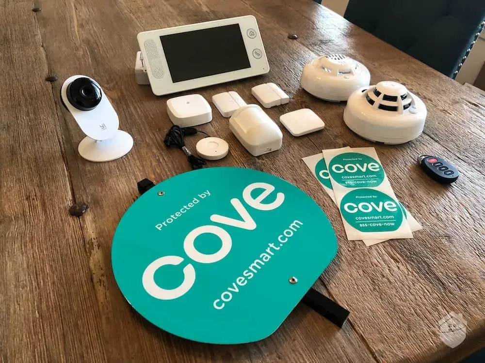 A variety of Cove home security devices including a camera, sensors, smoke detector, control panel, remote, and signage.