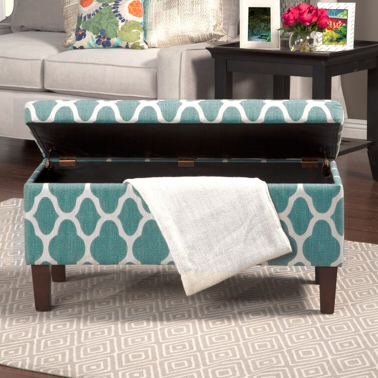 A teal and white patterned storage ottoman with an open lid and a white throw blanket draped over one corner.
