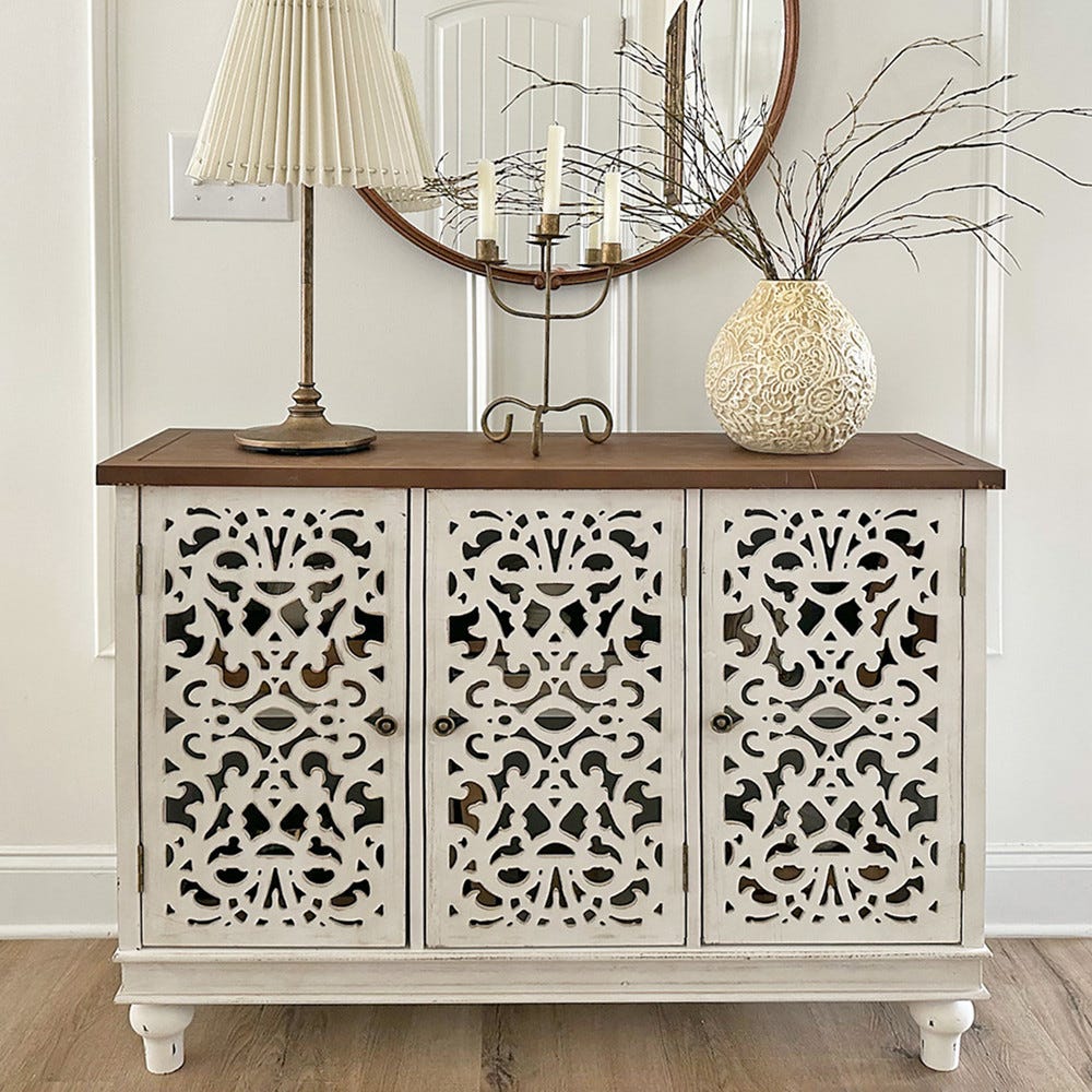 A wooden sideboard with ornate white panels, a table lamp, a round mirror, a candle holder, and a decorative vase.