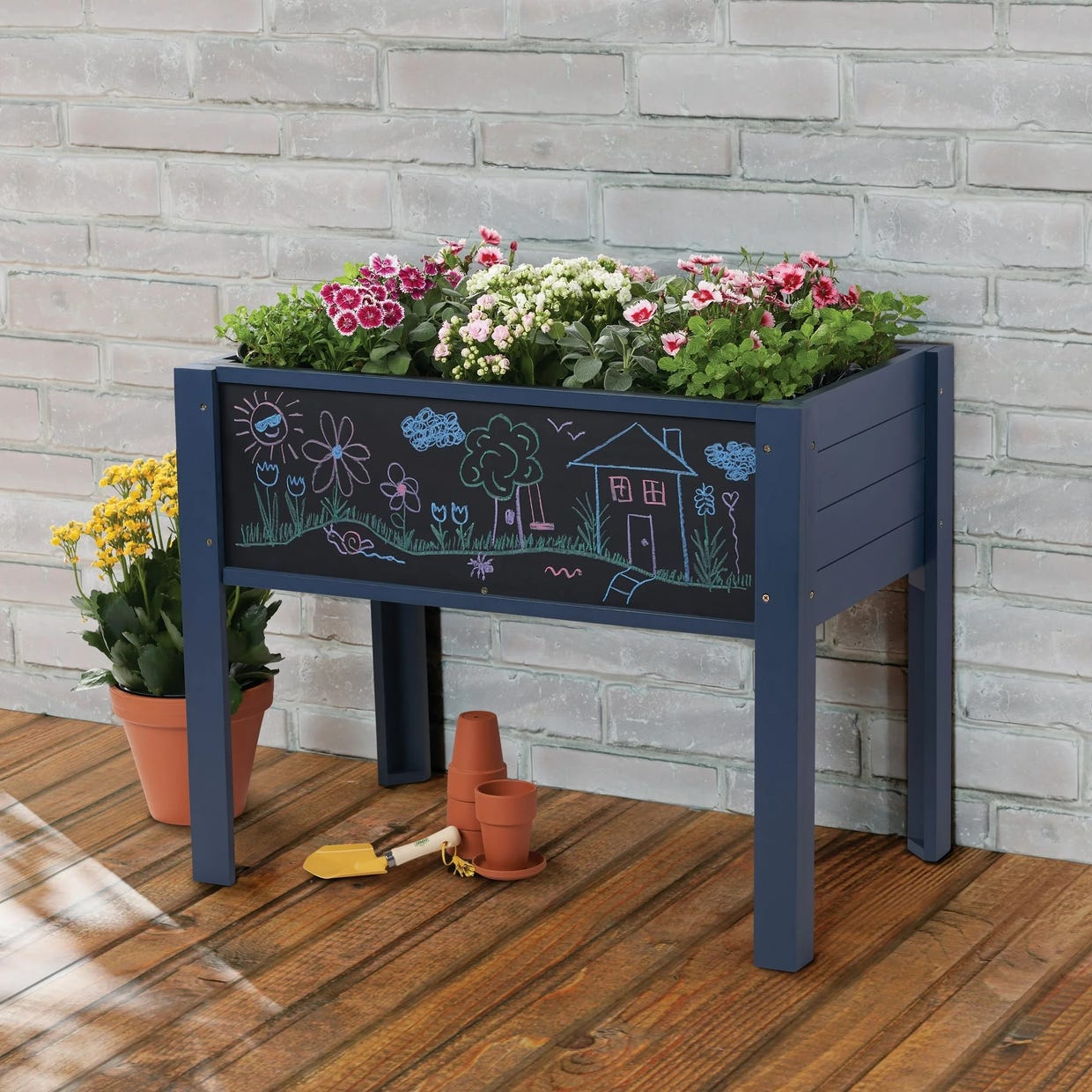 A raised garden bed with a chalkboard panel filled with plants on a wooden deck, a watering can, terracotta pots, and gardening tools beside it.