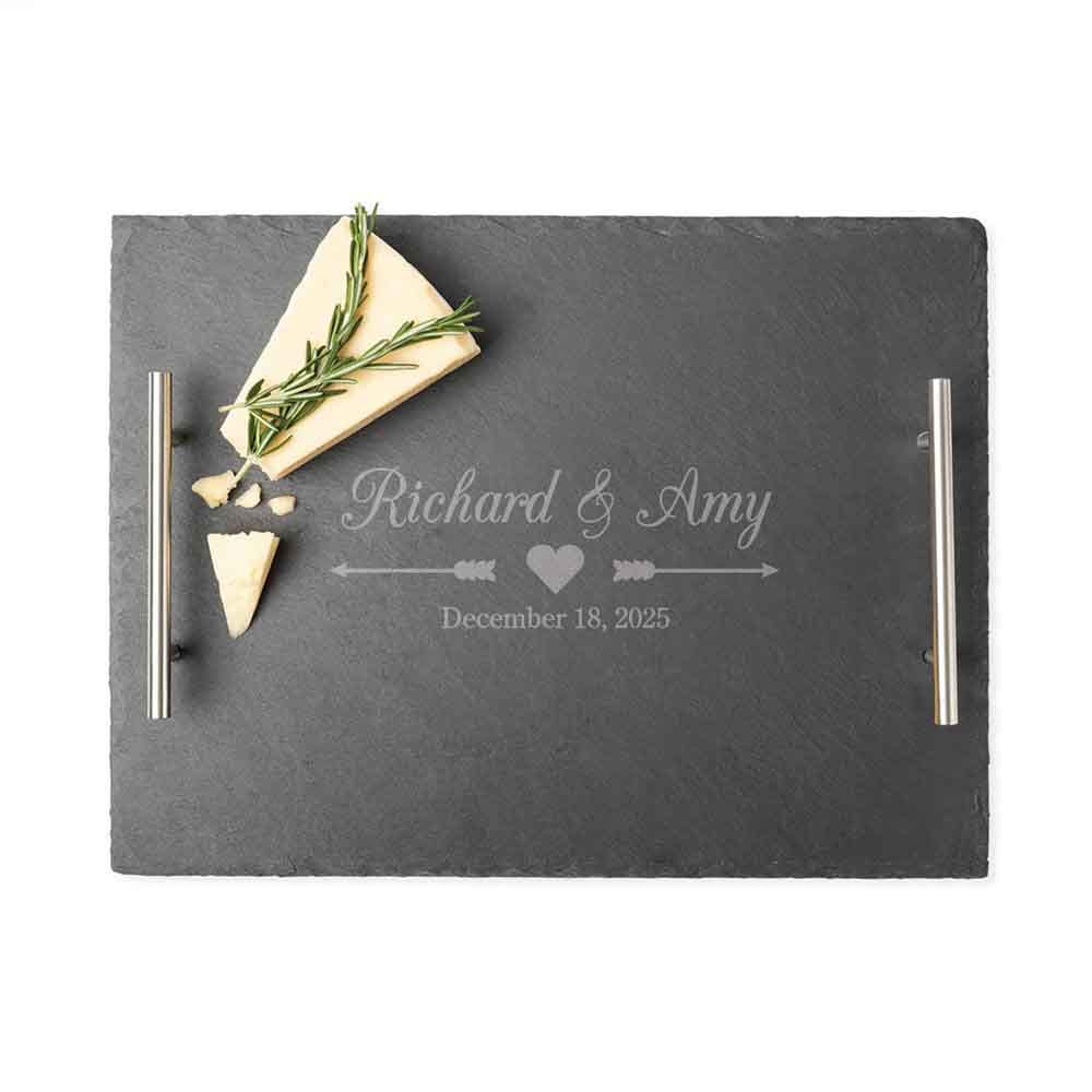 A black slate tray with metal handles features the personalized engraving \“Richard & Amy\“ above a heart and arrow design, along with the date \“December 18, 2025.\“ It includes a piece of cheese garnished with rosemary.