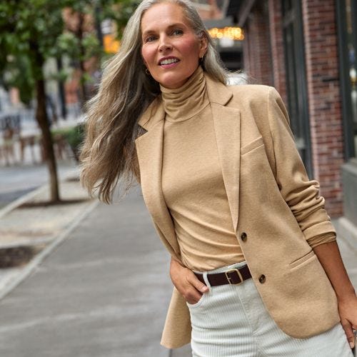 A woman is wearing a beige turtleneck top that fits close to the body with a high neck collar, paired with a tan blazer.