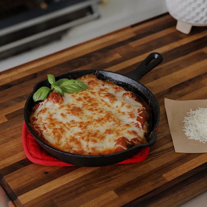 A cast-iron skillet containing a baked cheesy dish garnished with basil leaves on a wooden countertop.