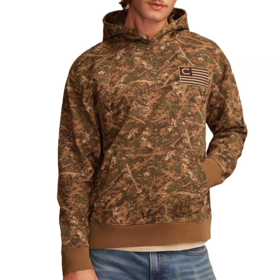 A camo-patterned hoodie featuring an American flag design with a \“C\“ emblem on the chest, with ribbed cuffs and hem, in earth tones. Paired with blue jeans.