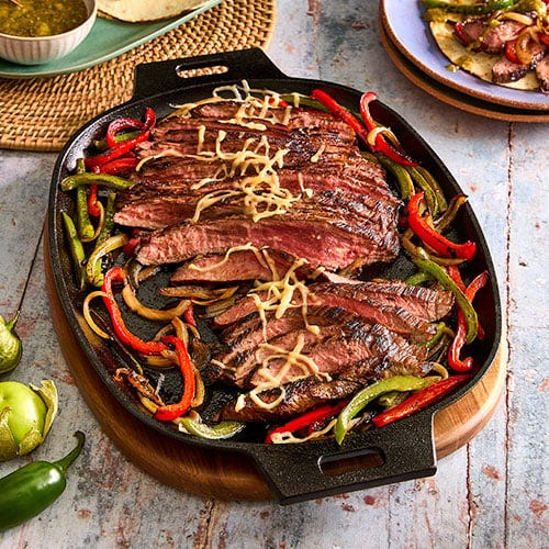 A cast iron skillet contains sliced steak and bell peppers, resting on a round wooden trivet.