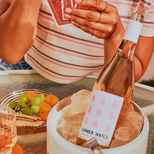 A bottle of Summer Water rosé wine in an ice bucket, with a person holding playing cards and a bowl of fruit in the background.