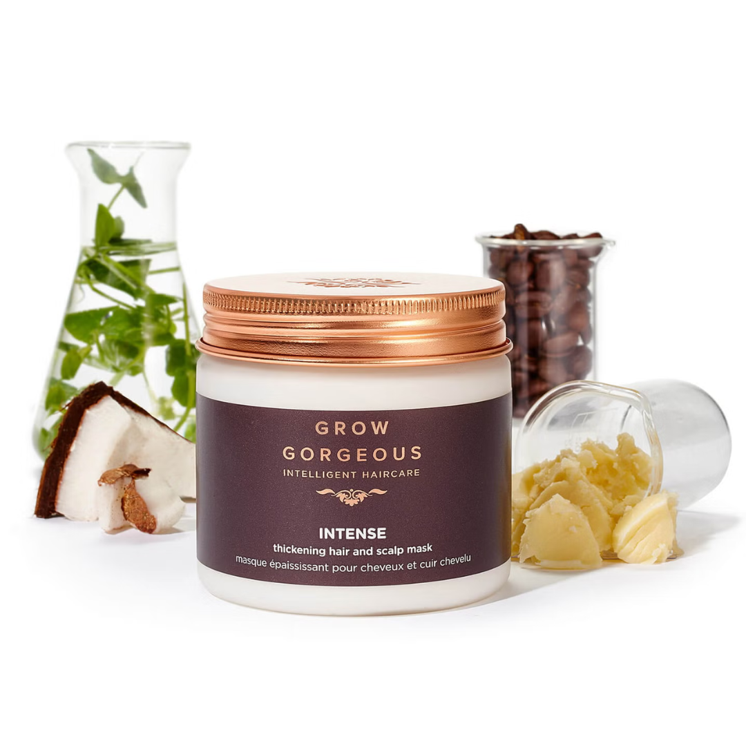 A jar of Grow Gorgeous hair and scalp mask, flanked by a coconut half and a laboratory flask with green leaves, and a spilled capsule with a yellowish substance.