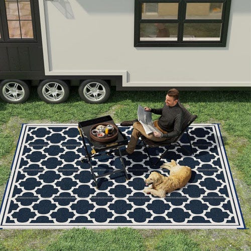 A man sitting on a folding chair on a patterned outdoor rug, with a portable grill and a lounging dog beside him.