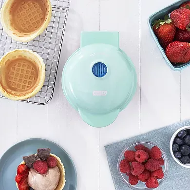 A mint green, circular Dash Mini Waffle Bowl Maker is shown alongside two waffle bowls and various berries.