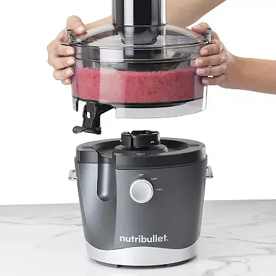 A NutriBullet Juicer with a person's hands locking a clear top onto the grey base; a crimson liquid is visible inside the machine.