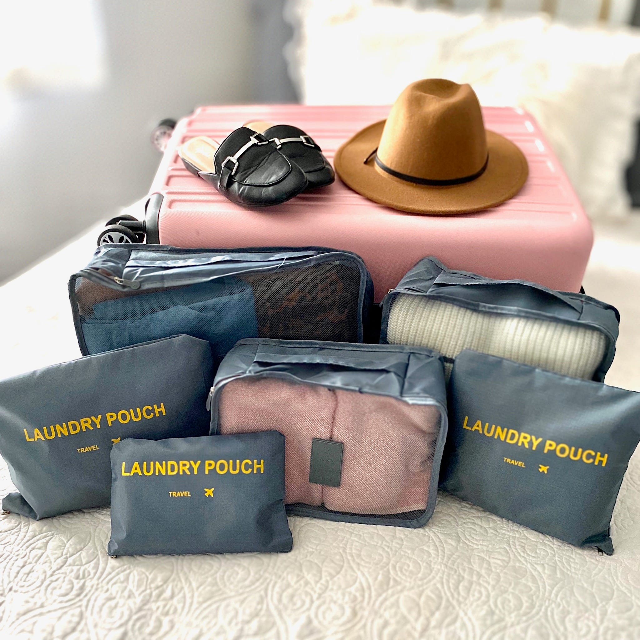 A pink suitcase, a brown hat, a black purse, and several blue packing cubes labeled 'LAUNDRY POUCH.'