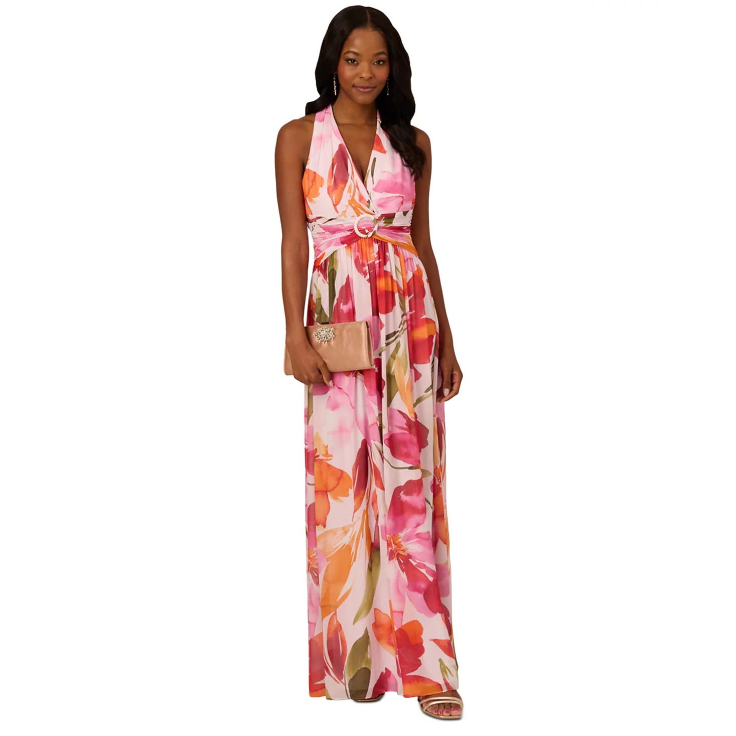 A woman is wearing a sleeveless, floral maxi dress with a deep V-neck and a pink belt. She is holding a beige clutch and wearing open-toe sandals.