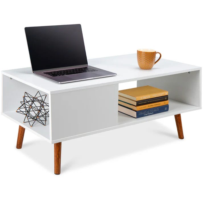 A white coffee table with a laptop, three books, a cup, and a decorative metal object on it, featuring angled wooden legs.
