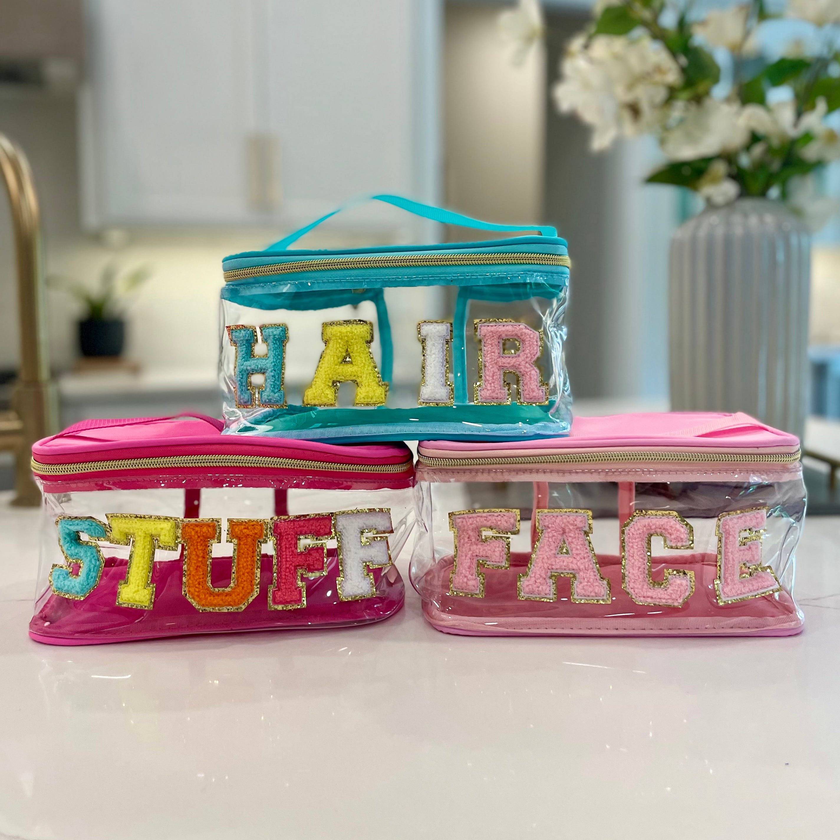 Three clear cosmetic bags with colorful letters on the front spell \“HAIR,\“ \“STUFF,\“ and \“FACE.\“ The bags are transparent with pink and teal accents, ideal for organizing personal care items.