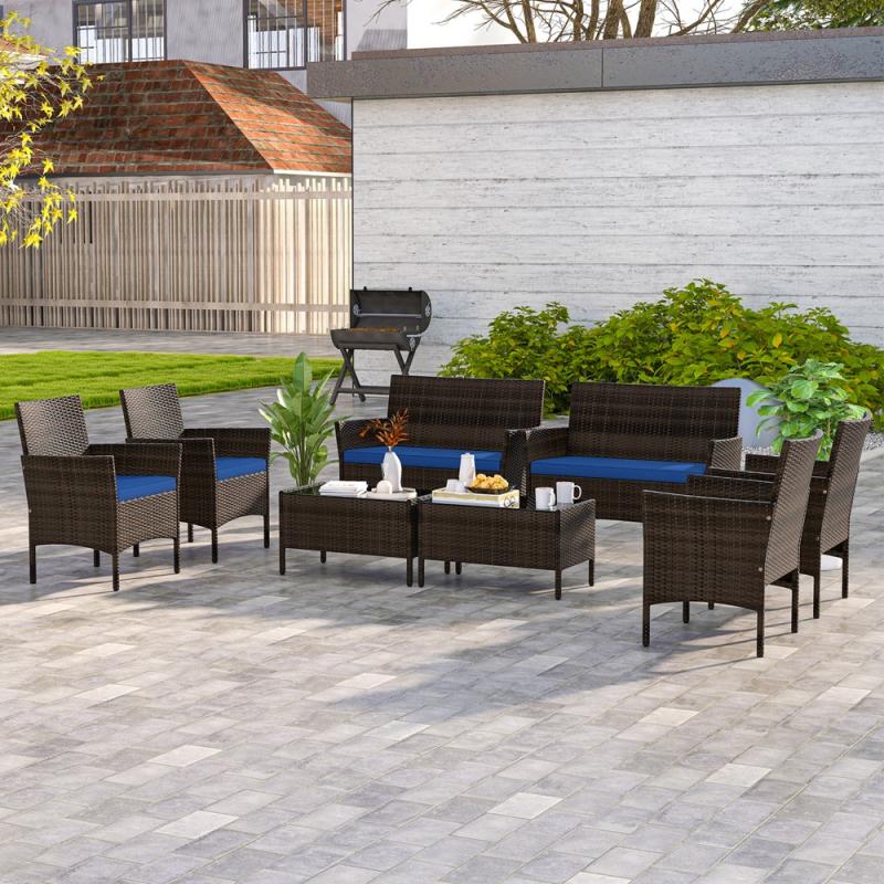 8-piece Costway patio set in navy includes two loveseats, four armchairs, and two coffee tables. The furniture features dark wicker with navy cushions, suitable for outdoor use, arranged on a tiled patio with a background of greenery and a grill.