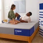A man and woman sitting on a Casper One mattress on a wooden bed frame, in a bedroom setting.