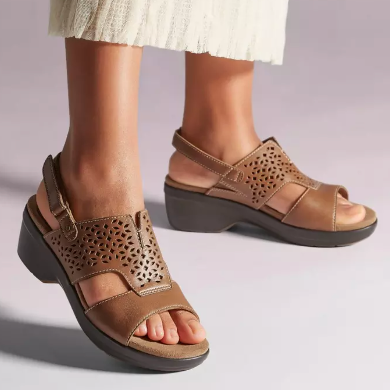 A pair of tan leather sandals with decorative cutouts and Velcro straps, worn by a person in a white skirt.